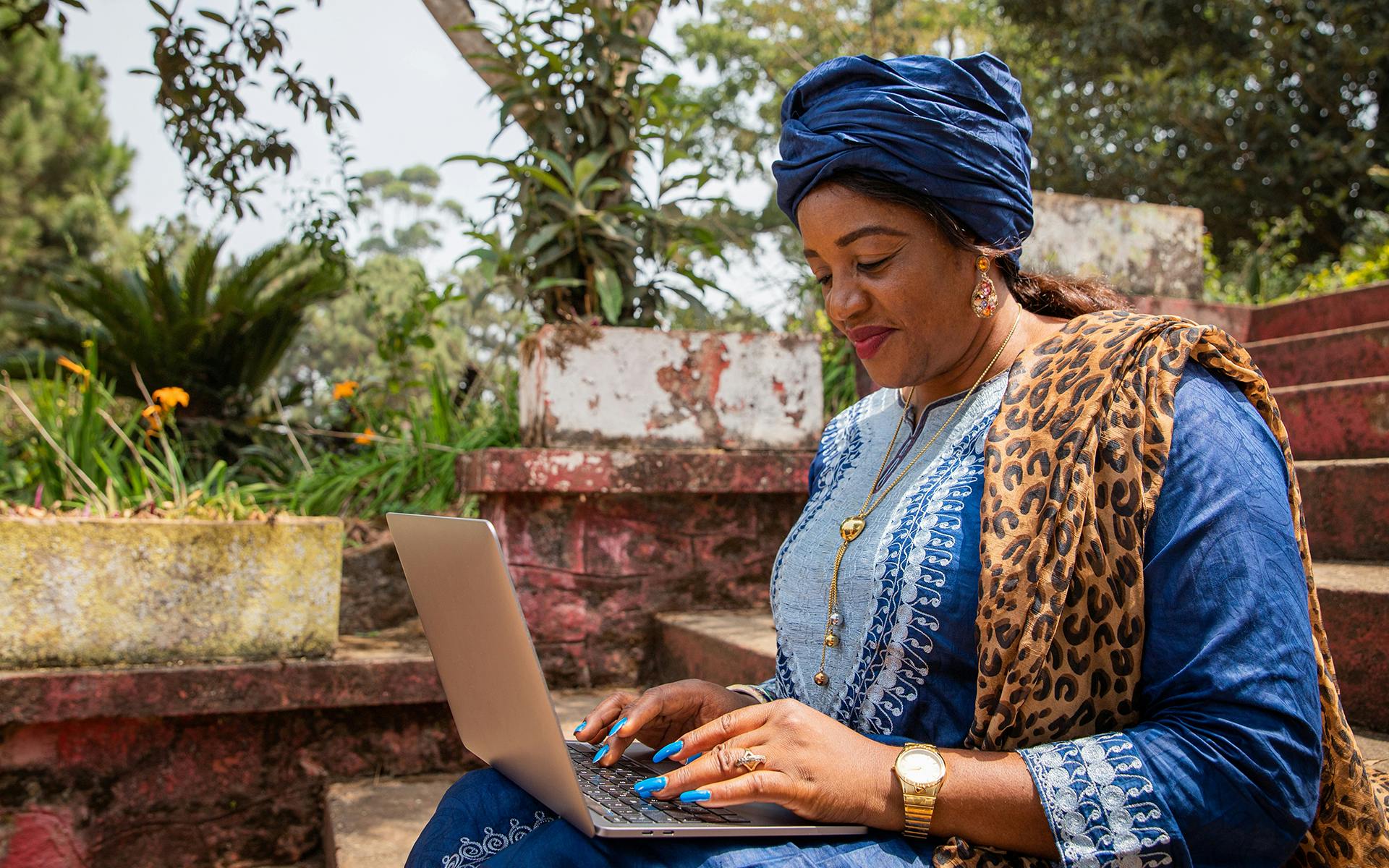 A middle-age African woman wearing a blue traditional dress and head wrap works on a computer.