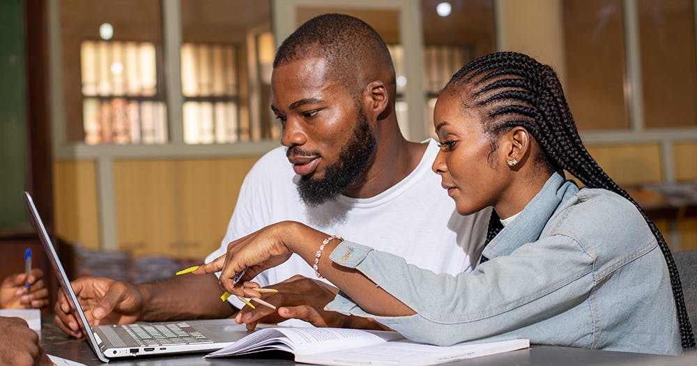 Two African university students--a woman with long braided hair and a man with short hair and a beard --look at a laptop while studying.